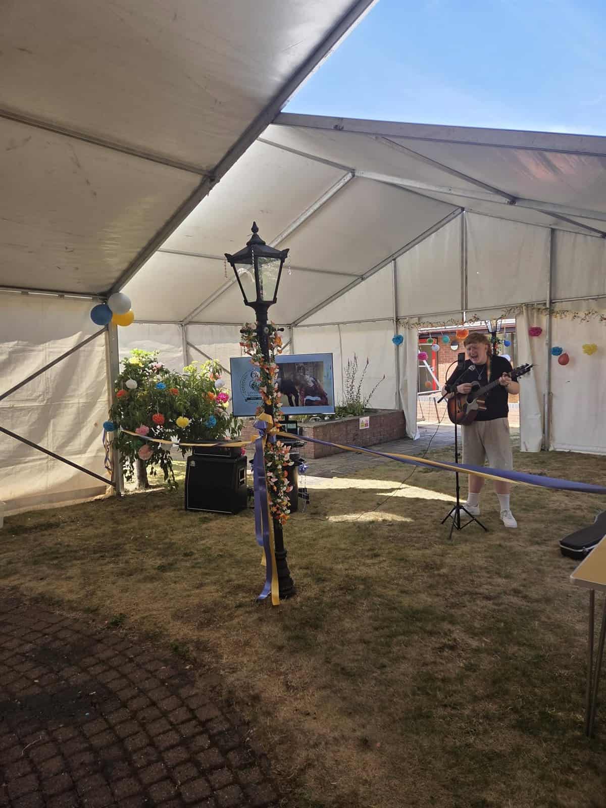 Alfie Kelly singing and playing guitar at an outdoor marquee event on a sunny day, decorated with flowers and balloons