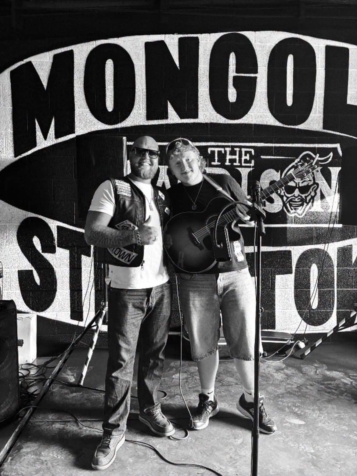Black and white photo of Alfie Kelly holding his guitar beside a supporter giving a thumbs up, in front of a large painted Steel Town mural